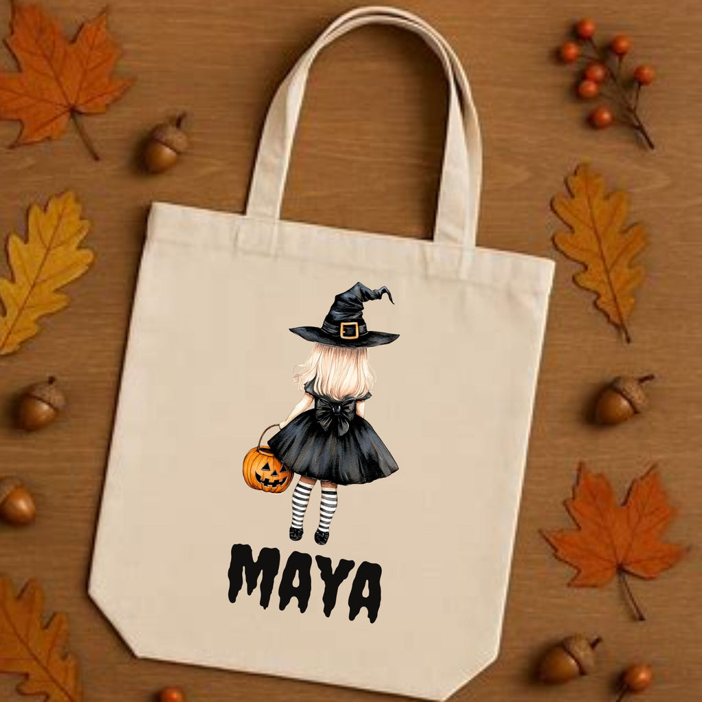 A cream canvas tote bag with a cartoon witch girl graphic and the name 'MAYA' printed in black, displayed on a wooden surface surrounded by autumn leaves, acorns, and small orange berries.