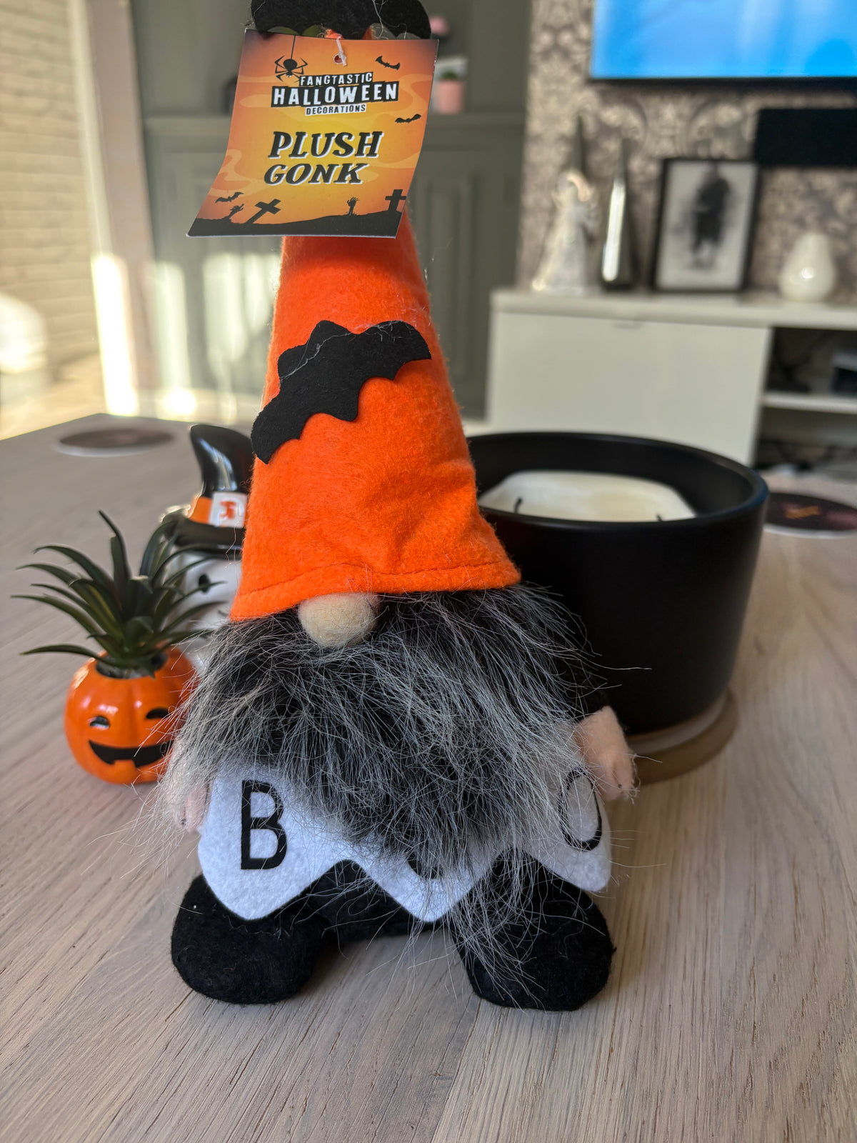 A Halloween-themed plush gnome figurine sitting on a light wood table. The gnome has an orange pointed hat with a black bat detail, a gray beard, and a white tattered shirt with black letters.