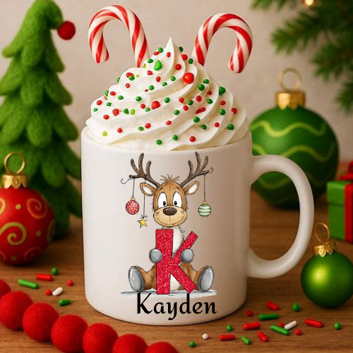 A white ceramic mug filled with whipped cream and topped with two candy canes, surrounded by Christmas decorations including ornaments, a tree, and sprinkles. The mug is personalized with a cartoon reindeer, a large glittery red letter 'K', and the name 'Kayden'.