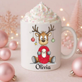 A white ceramic mug topped with whipped cream and sprinkles, featuring a cartoon reindeer wearing a red sweater and the name 'Olivia' printed below. The mug is styled with pink ornaments and warm fairy lights in the background.