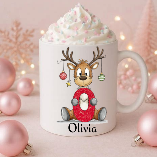 A white ceramic mug topped with whipped cream and sprinkles, featuring a cartoon reindeer wearing a red sweater and the name 'Olivia' printed below. The mug is styled with pink ornaments and warm fairy lights in the background.