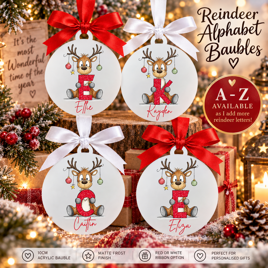 Four personalized round acrylic Christmas baubles displayed on a rustic wood stack with holiday decorations. Each bauble features a cartoon reindeer holding a letter in red glitter, with red and white ribbons.