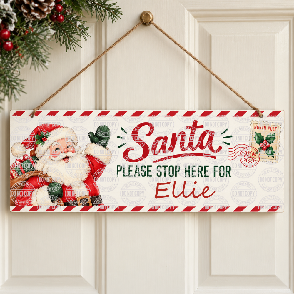A rectangular Christmas hanging sign with a red-and-white striped border, a vintage Santa illustration on the left, and the text 'Santa PLEASE STOP HERE FOR Ellie' in red and green lettering. The sign hangs from a rope on a white paneled door, with a holiday wreath visible in the upper left corner.