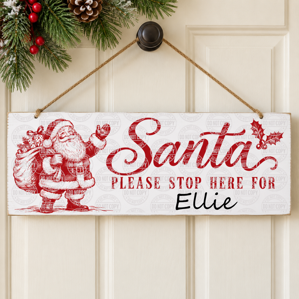 A rectangular white hanging sign with a red Santa illustration and the text 'Santa PLEASE STOP HERE FOR Ellie', hung on a cream paneled door with a wreath above.