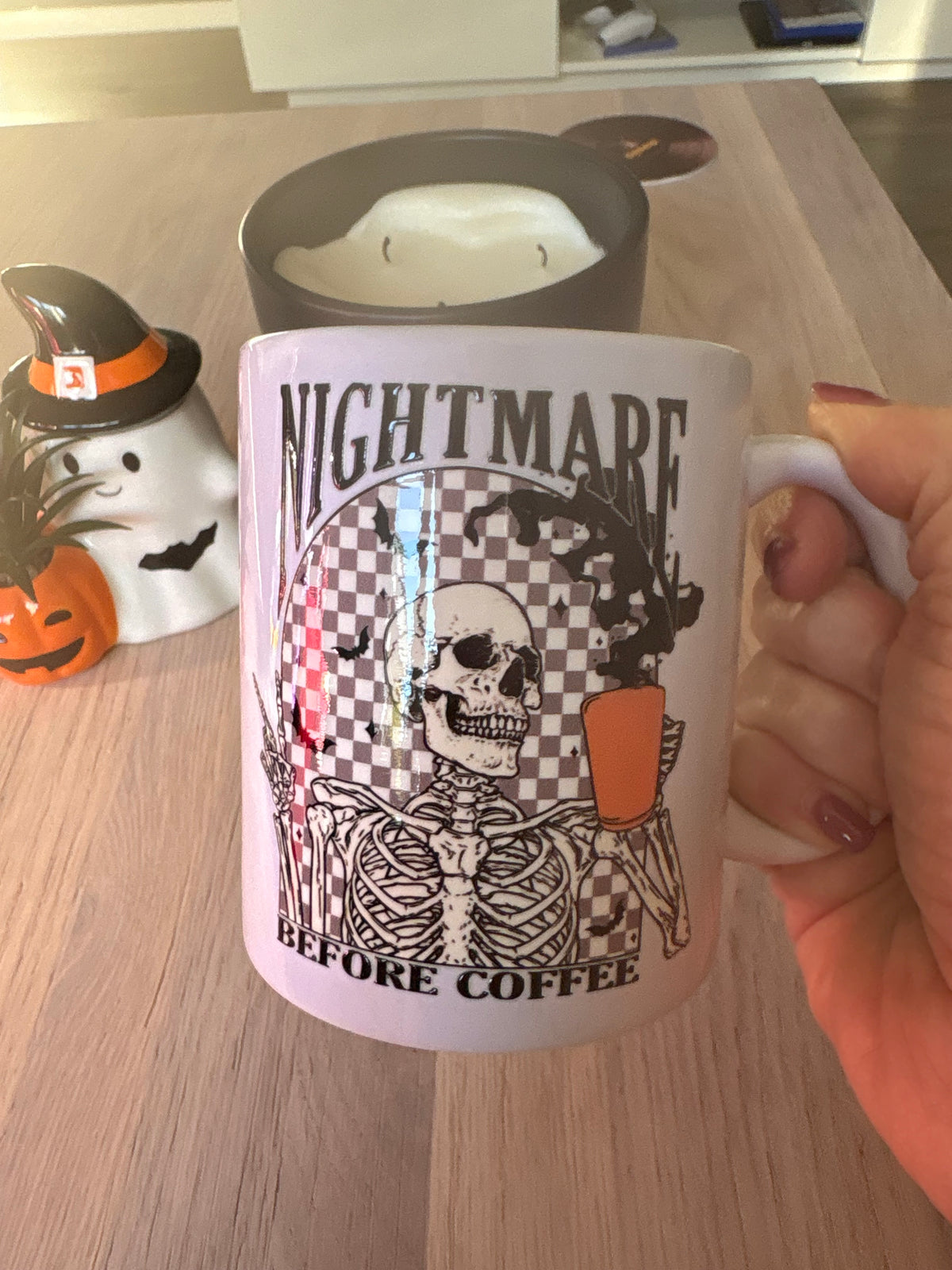 A hand holds a white ceramic mug with a Halloween-themed graphic that reads 'NIGHTMARE BEFORE COFFEE' featuring a skeleton holding an orange mug. The background shows a wooden table with other home decor items.