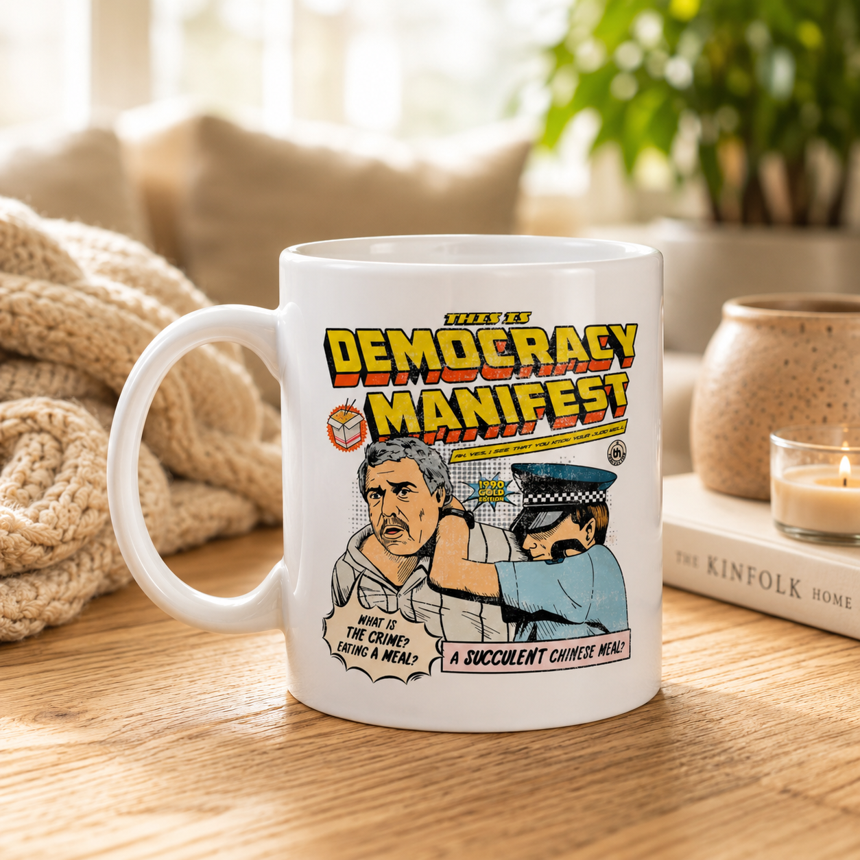 A white ceramic mug with a colorful comic-style graphic reading 'Team of Democracy Manifest' and an illustration of two people, placed on a wooden table in a cozy indoor setting with a blanket, book, and candle in the background.