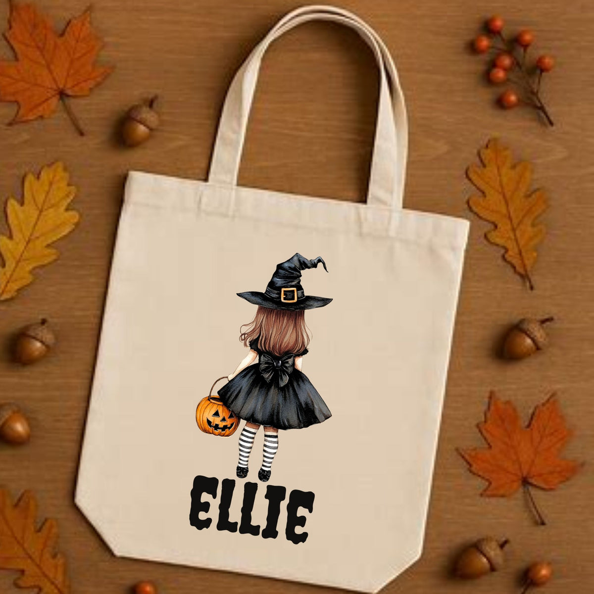 A cream canvas tote bag with a Halloween-themed illustration of a girl in a witch hat holding a pumpkin, with the name 'ELLIE' printed below in black. The bag is displayed on a wooden surface surrounded by autumn leaves, acorns, and small orange berries.