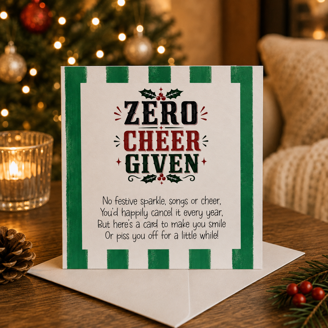 A Christmas greeting card with a white background, green striped border, and bold text reading 'ZERO CHEER GIVEN' with a humorous poem below, displayed on a wooden table with a candle, pinecone, and blurred Christmas tree in the background.