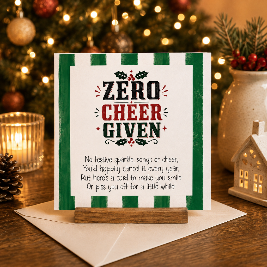 Zero Cheer Given funny cheeky Christmas card with green stripe border and holly illustration, standing on a wooden card holder next to a white envelope