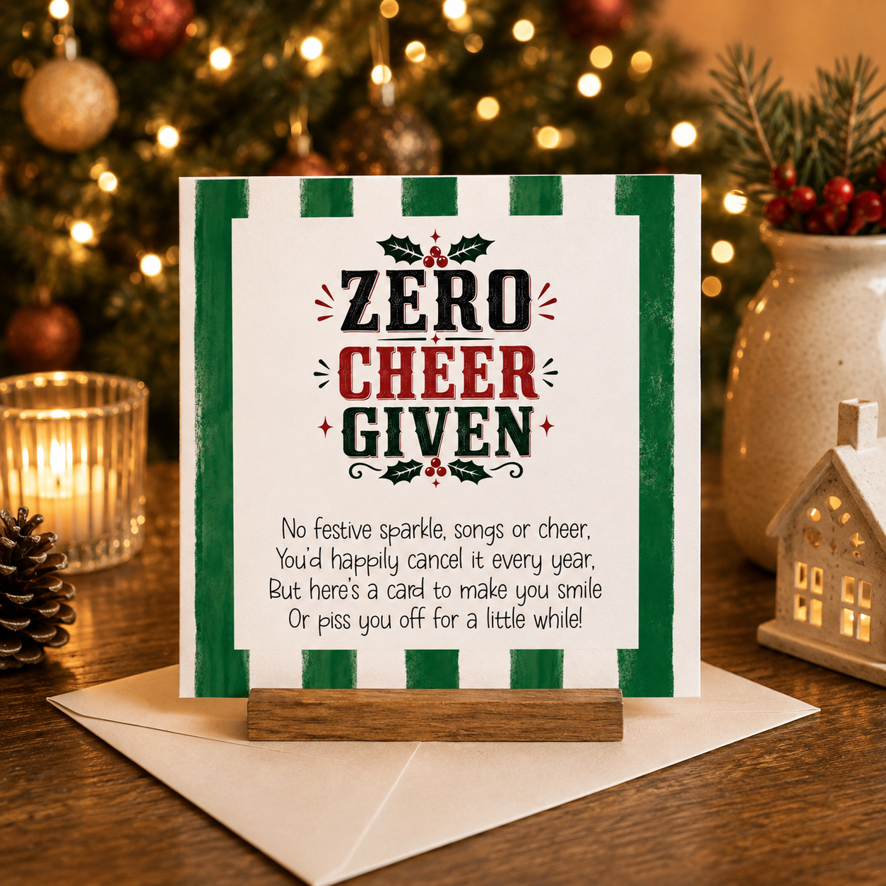 Zero Cheer Given funny cheeky Christmas card with green stripe border and holly illustration, standing on a wooden card holder next to a white envelope